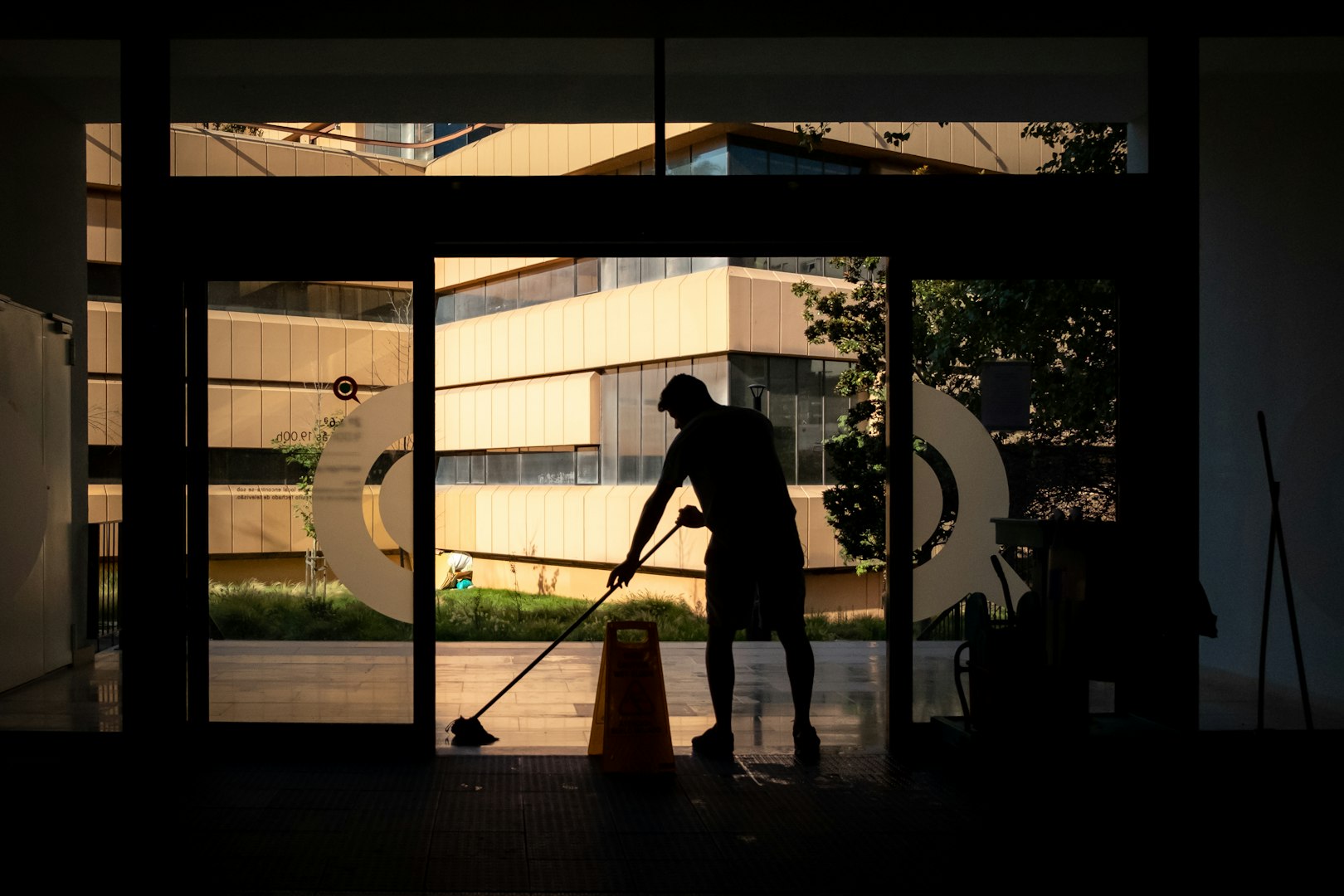 A woman with long hair, wearing a white t-shirt and yellow cleaning gloves, is cleaning a window with a cloth. Sunlight streams through the window, illuminating her smile and the indoor plants nearby.